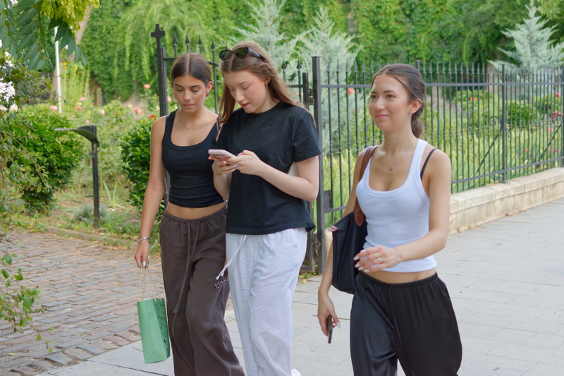 19 - Three young people walking together on a sidewalk next to a metal fence of a mini park
